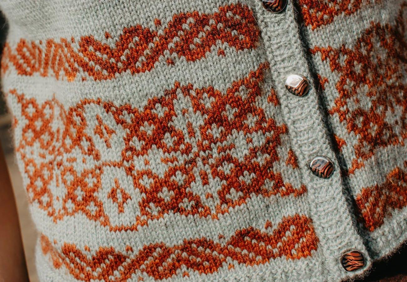 Close-up of the Ailis Sweater showing a burnt-orange Latvian-inspired colourwork motif on a pale grey-green body with decorative buttons