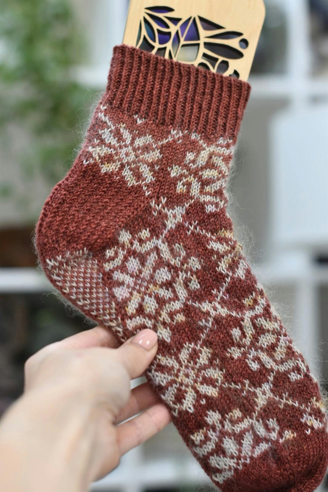 A toe-up colourwork sock in rust red with cream snowflake motifs held on a wooden sock blocker